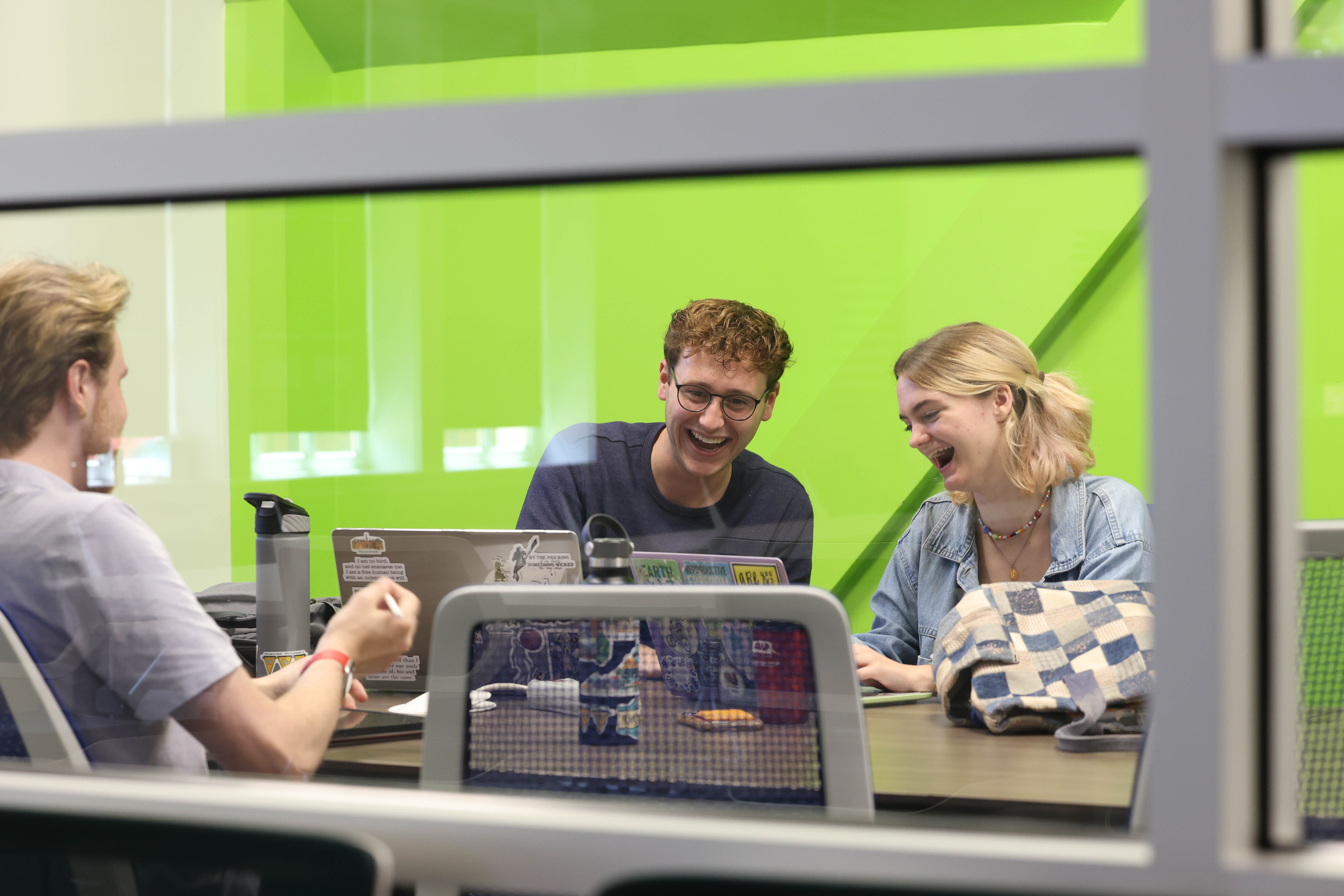 Three students smile looking at a computer in a bright green collaborative space