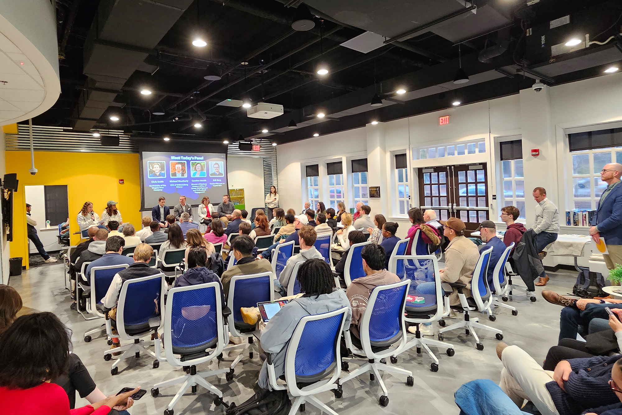 A room full of seated event attendees and a panel of speakers